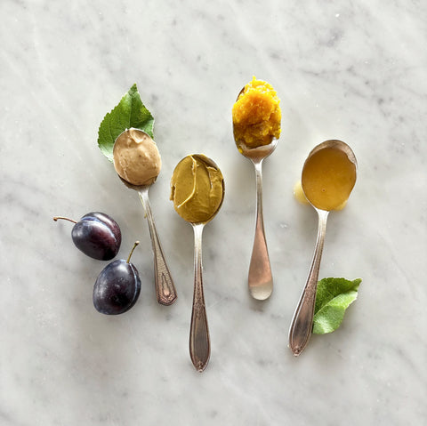Four silver spoons filled with almond cream, pistachio cream, Sicilian orange marmalade and orange blossom honey, arranged on a light marble surface with two plums and green leaves.