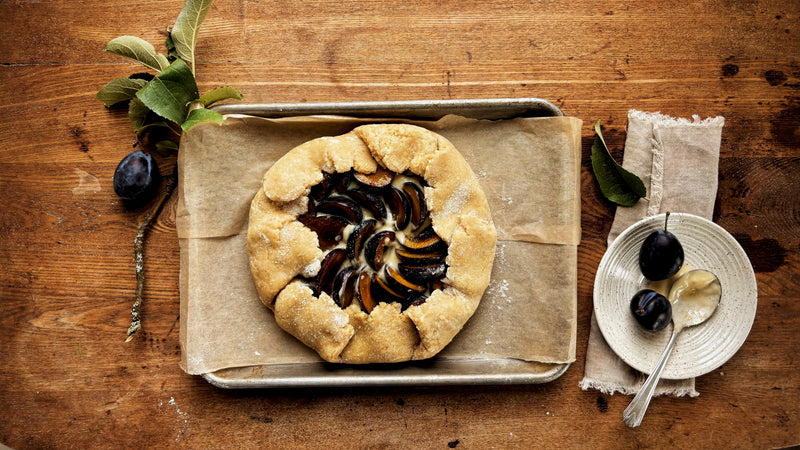 Baked plum and almond crostata on parchment on a sheet pan, with fresh Italian plums, plum leaves and a small plate of plums on a dark wood table