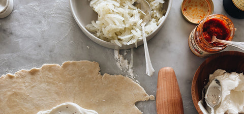 Rolled-out crostata dough on a marble counter with ricotta, yellow squash and onion piled in the center, beside a wooden rolling pin, a bowl of ricotta and a jar of Tutto Sicilia Hot Pepper Cream.