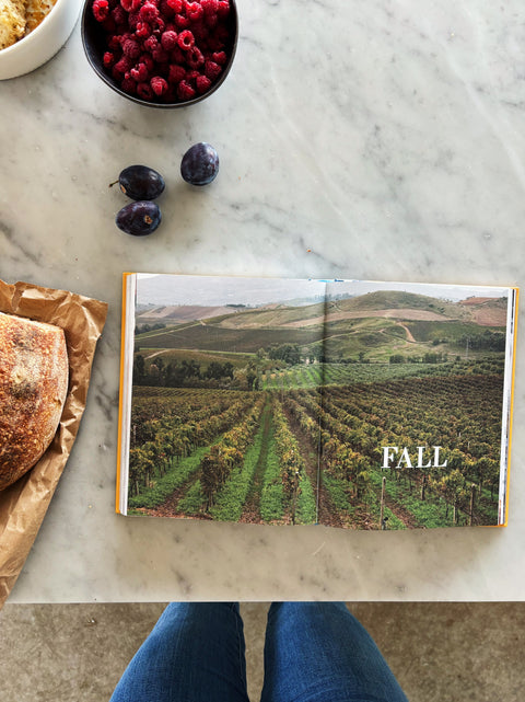 An open cookbook spread on a white marble surface, showing a vineyard landscape with the word 'FALL' on the page. A bowl of raspberries, three plums, a loaf of bread on brown paper, and a woman's blue jeans are visible around it.